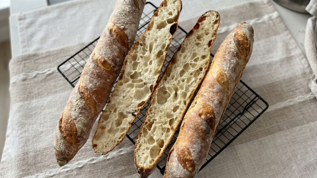 Three golden sourdough baguettes with scored tops cooling on a wire rack
