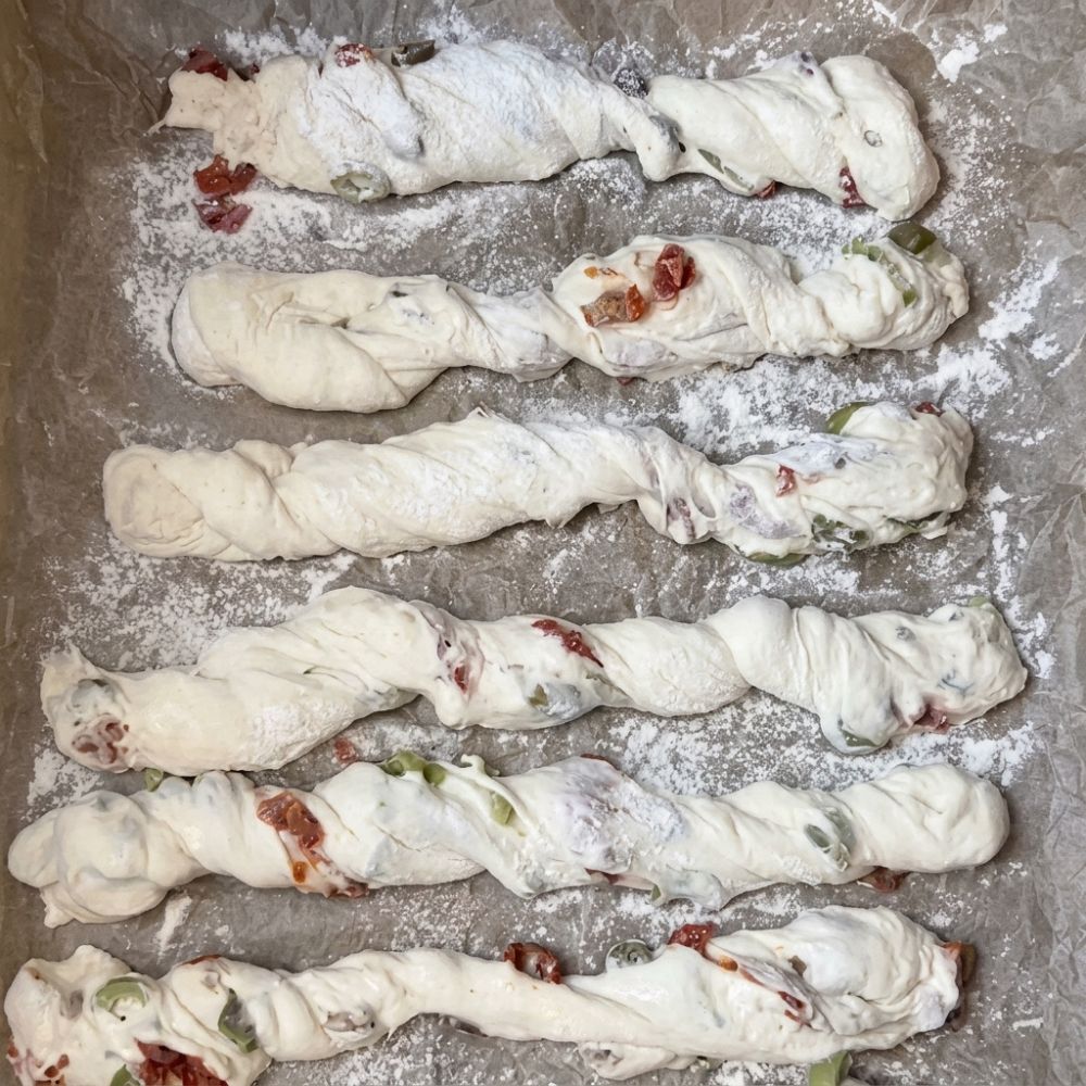 Twisted mediterranean sourdough breadsticks proofing on a floured tray before baking